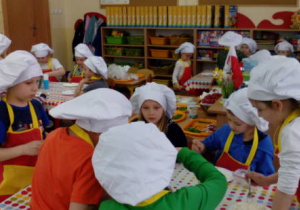 Ujęcie przedstawia salę przedszkolną, a w niej dzieci przy stolikach podczas zajęć kulinarnych.